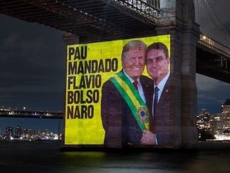 Projeção em NY mostra Flávio entregando faixa a Trump: "pau mandado". - Reprodução/YouTube